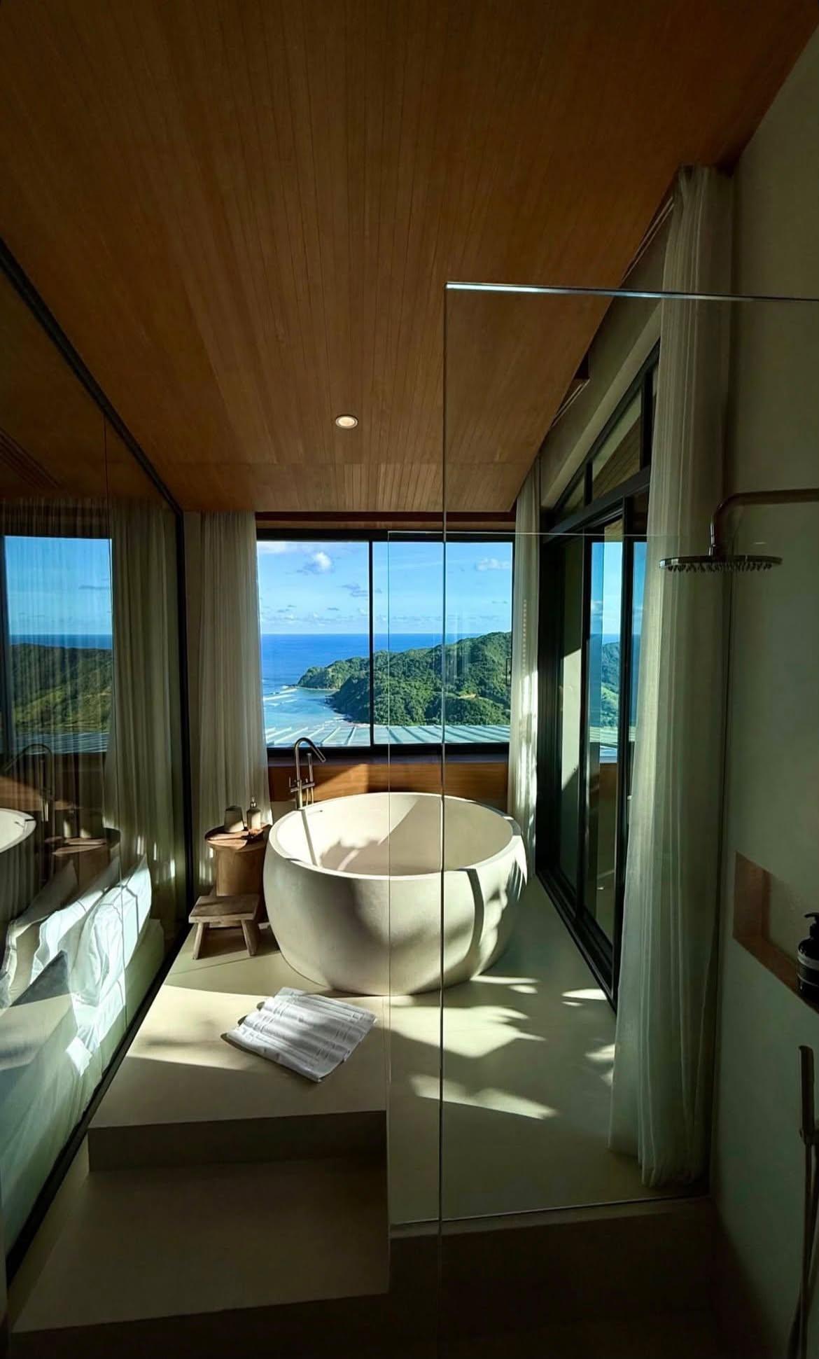 A sculptural stone bathtub facing floor-to-ceiling views of the ocean and green hills